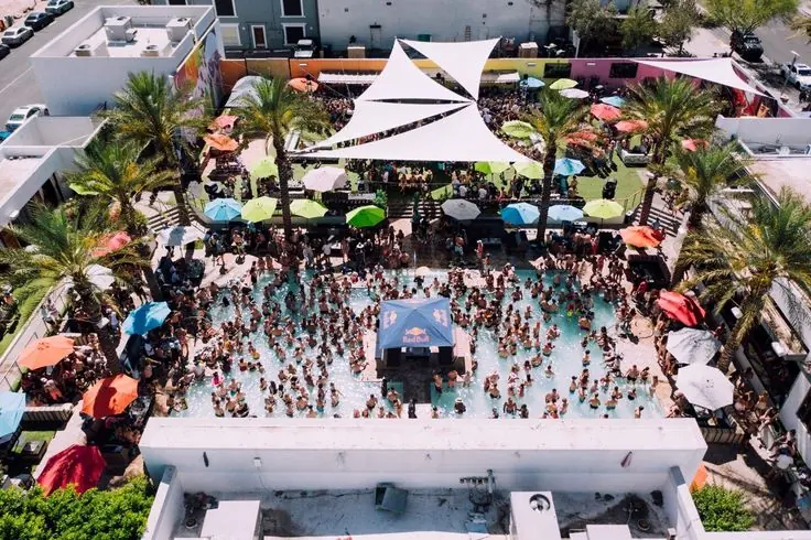 Aerial view of a dayclub pool ringed by palms, umbrellas and cabanas
